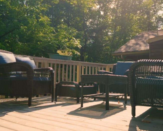 The image shows a set of woven patio furniture arranged on a wooden deck surrounded by trees.