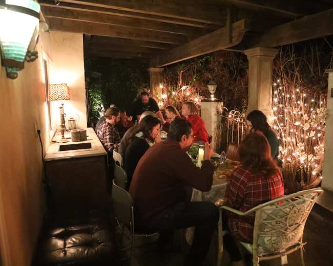 A group of people is seated around a table on a patio decorated with string lights and surrounded by a garden.
