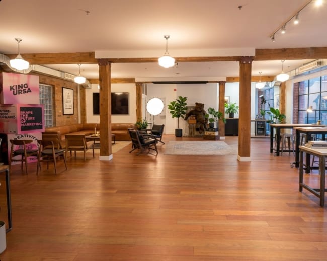 The image shows a spacious, well-lit office space with wooden floors, decorative potted plants, and seating areas arranged around a central gathering space.
