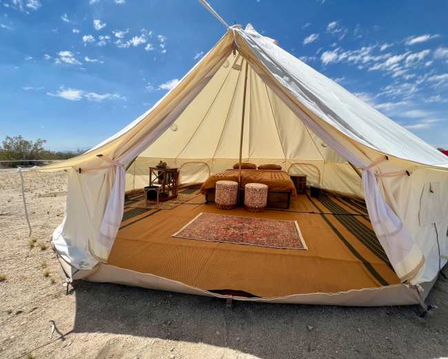 A spacious, cream-colored canvas tent is set up in a desert landscape, featuring a neatly arranged interior with a bed, two poufs, and a decorative rug.