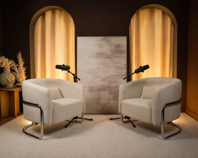 The image shows a modern, minimalistic interview setup with two beige armchairs facing each other, a neutral backdrop, and two microphones on stands positioned between them.