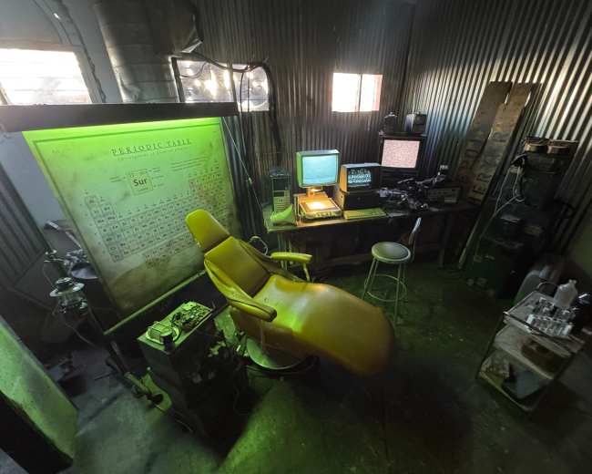 The image shows a dimly lit room with a yellow dental chair, a vintage computer setup, and various equipment against a corrugated metal wall.
