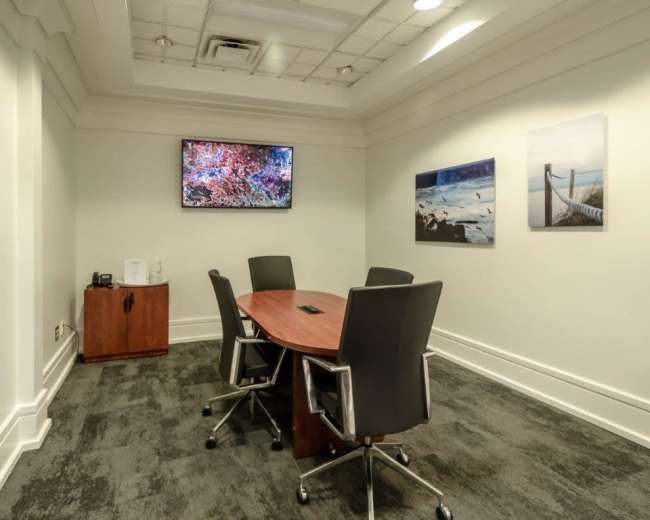 The image shows a small conference room with a wooden table and four black chairs, featuring a TV mounted on the wall and two framed pictures.