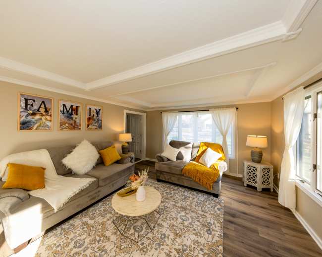 The living room features a beige sectional sofa with decorative pillows, a warm throw blanket, a round coffee table, and large windows allowing natural light, complemented by wall art spelling "FAMILY."