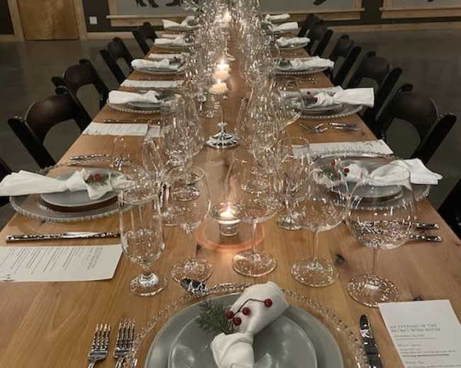 A long wooden dining table is elegantly set with fine china, glassware, and white napkins, surrounded by chairs in a room featuring silhouette artwork on the walls.