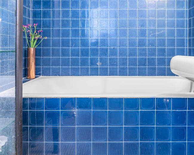 A blue-tiled bathroom features a white bathtub and a small vase with flowers on the edge.