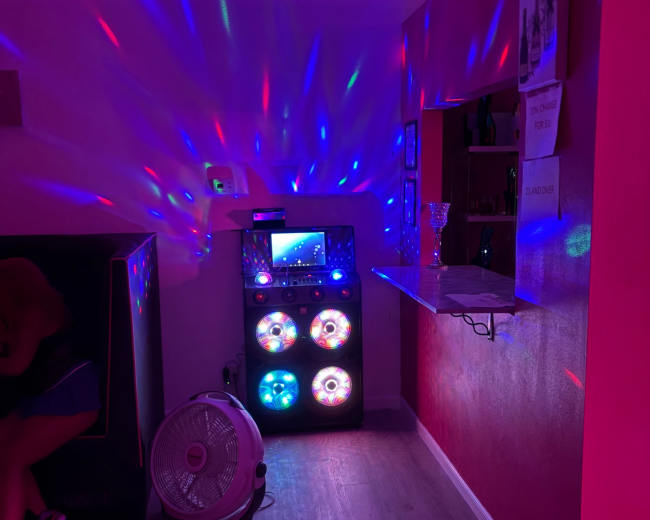 The image shows a brightly lit room with colorful lights creating patterns on the walls, featuring a sound system with colorful speakers and a fan in the corner.