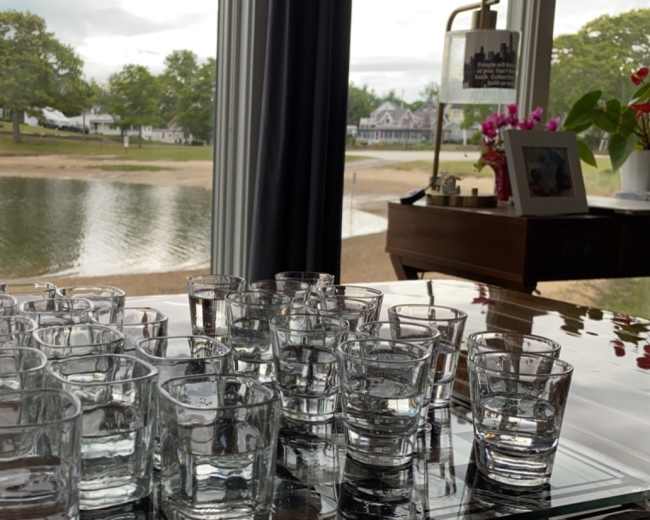 A row of empty glass shot glasses is arranged on a tray near a window overlooking a calm water view.
