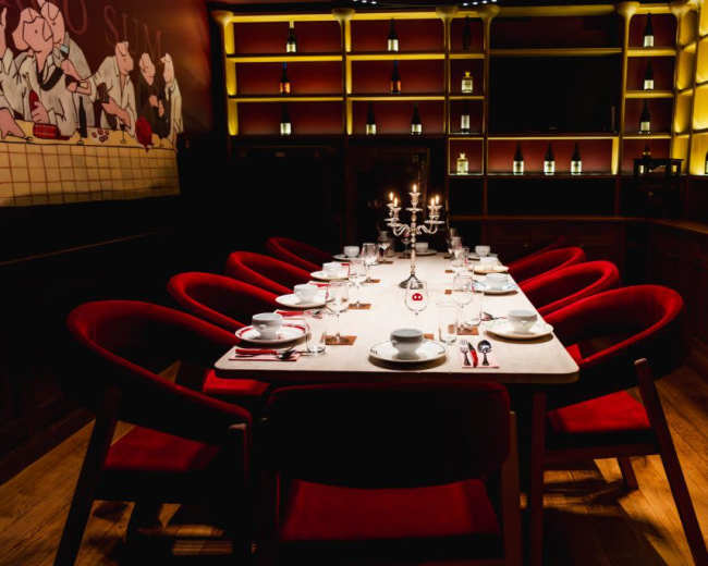 A long dining table with red chairs is centrally located in a dimly lit room, surrounded by shelves displaying bottles.