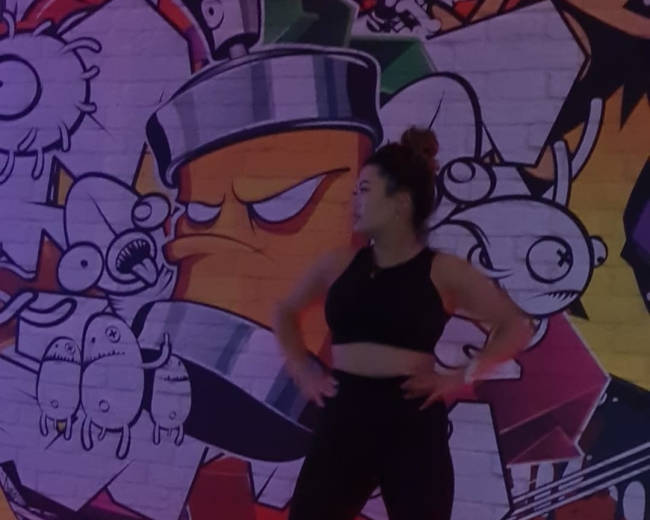 A woman wearing a crop top and black leggings performs on roller skates against a vibrant graffiti mural with colorful cartoon characters.