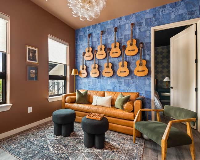 The image features a cozy room with a brown wall, a blue accent wall adorned with guitars, a leather sofa, two small black stools, and a green armchair.