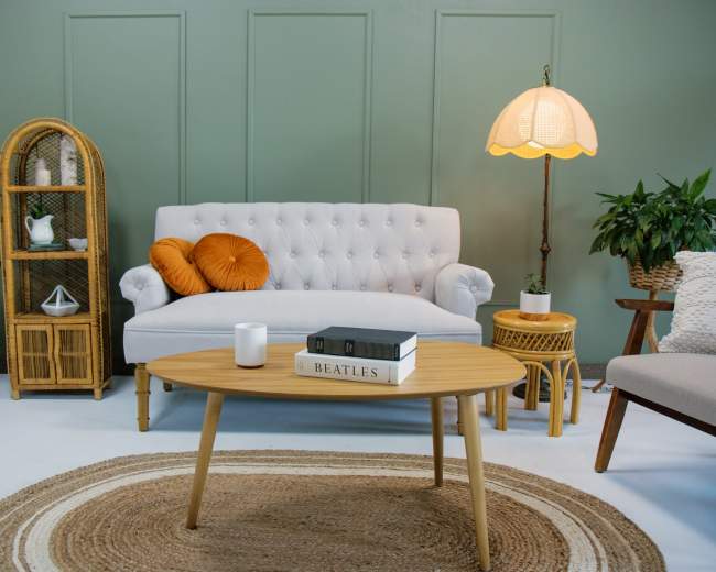 A light grey tufted sofa sits in a green room alongside a round woven rug, a wooden coffee table with books, and decorative plants.