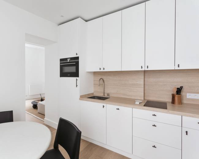 A modern kitchen features white cabinets, a beige countertop, a stainless steel sink, and a round table with black chairs.