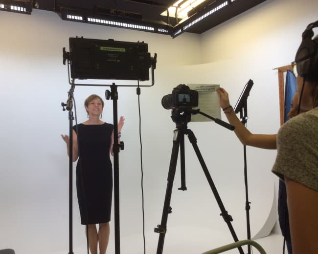 A woman stands in front of a camera and lights while holding a script, as another person operates the camera in a studio setting.