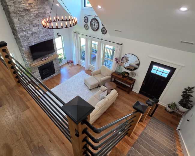 The image shows a spacious living room with a stone fireplace, large windows, and modern decor, viewed from a staircase railing.