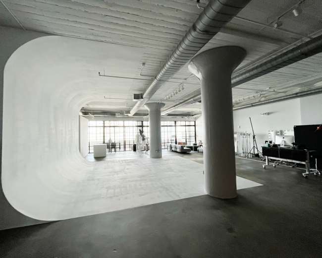 A spacious studio with a smooth, curved white backdrop, large windows allowing natural light, and various pieces of furniture and equipment.