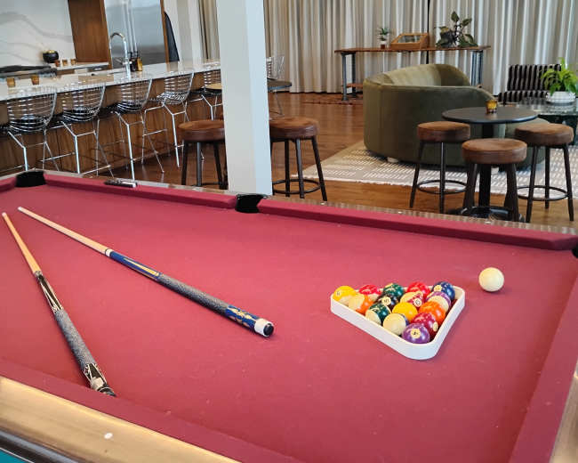 A billiard table with two cue sticks and a set of colorful pool balls is positioned in a modern recreational space featuring a kitchen and seating area in the background.