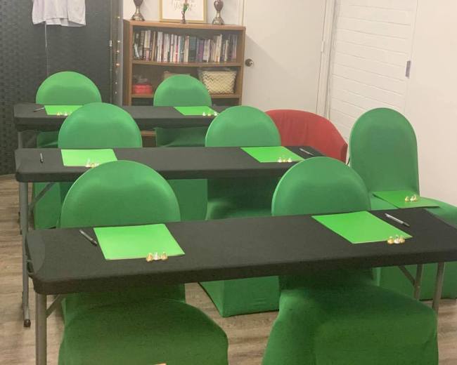 The image shows a room arranged with several green chairs and black tables, each equipped with a green notepad and a pen.
