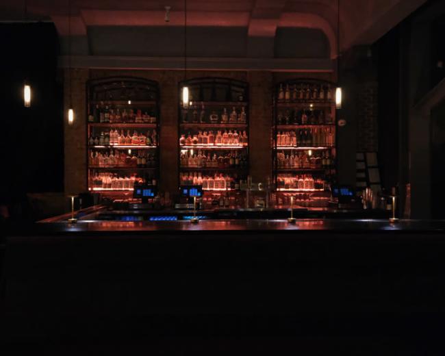 A dimly lit bar features a long counter with illuminated shelves filled with various bottles in the background.