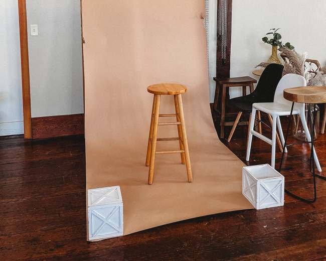 A photo studio setup with a wooden stool positioned in front of a tan backdrop and rolls of colored paper hanging above.