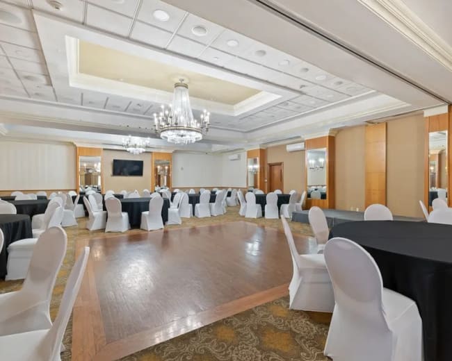A banquet hall with round tables covered in black and white tablecloths, surrounded by white chairs and illuminated by a chandelier.
