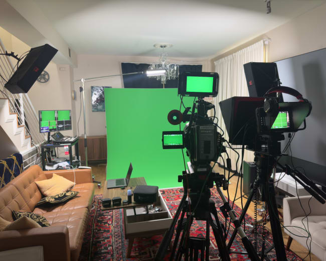 A studio setup features multiple cameras, lighting equipment, and a green screen, arranged in a well-furnished room.