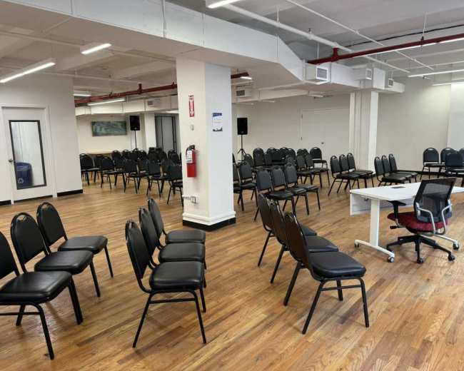 The image shows a large room filled with rows of black chairs arranged for an event, with a white table and a chair positioned toward the front.