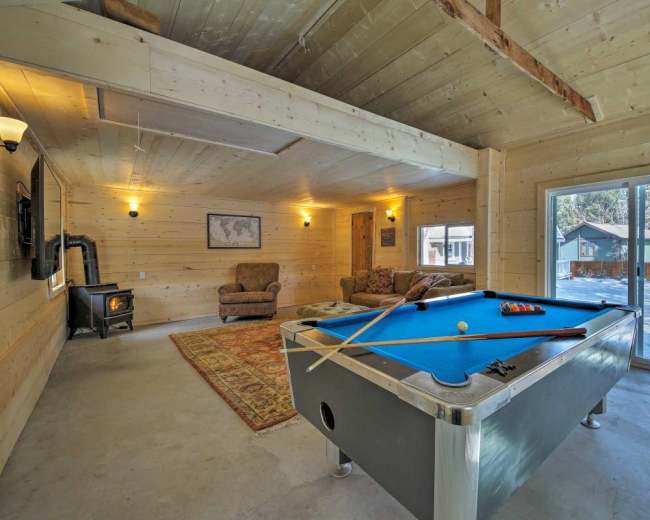 A light-colored wooden interior with a blue pool table, a sofa, and a wood stove, overlooking a snowy outdoor area through large glass doors.