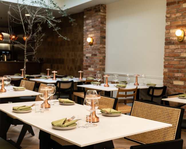 A modern dining space features tables set with green napkins, glassware, and copper accents, complemented by brick walls and soft lighting.