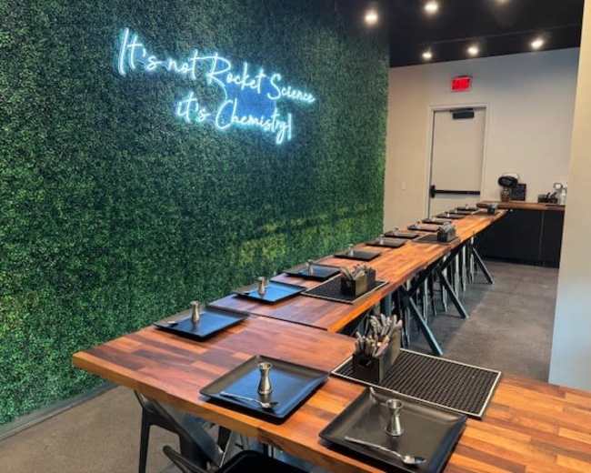 The image shows a modern dining area with a wooden table set for guests, featuring black plates, utensils, and a neon sign on a green wall that reads "It's not Rocket Science, It's Chemistry."