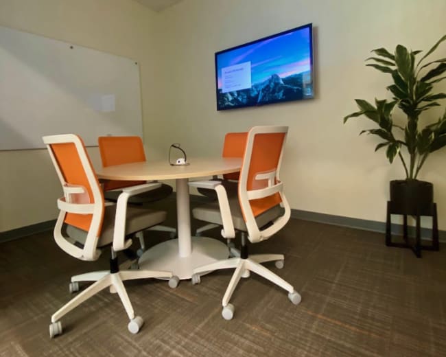 A small conference room features a round table surrounded by four chairs with orange cushions, a wall-mounted screen displaying a presentation, and a potted plant in the corner.