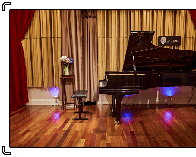 A black grand piano stands beside a stool on a polished wooden floor, with a vase of flowers on a nearby table and soundproof curtains in the background.