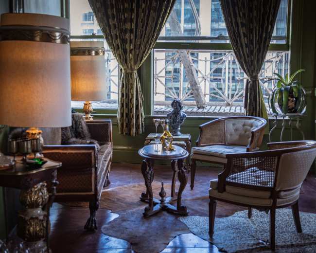 A cozy sitting area features vintage furniture, including two chairs, a table, and a decorative lamp, with sunlight streaming through a large window.