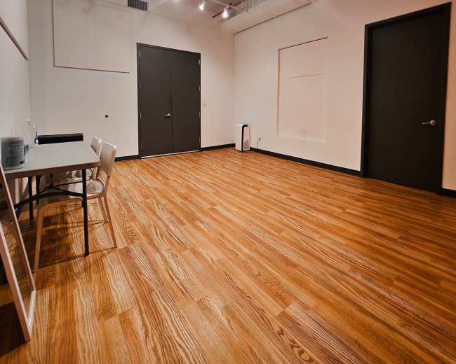 The image shows an empty room with wooden flooring, a table with chairs, and two closed doors.