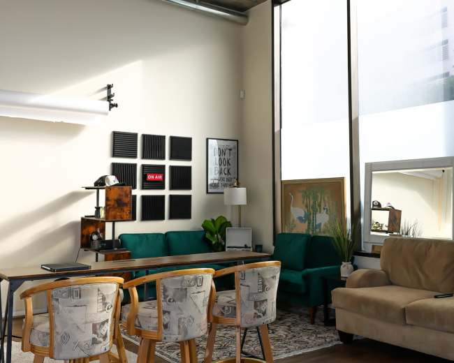 The image shows a modern living space featuring a green sofa, a beige couch, a dining area with high stools, and large windows allowing natural light to enter.