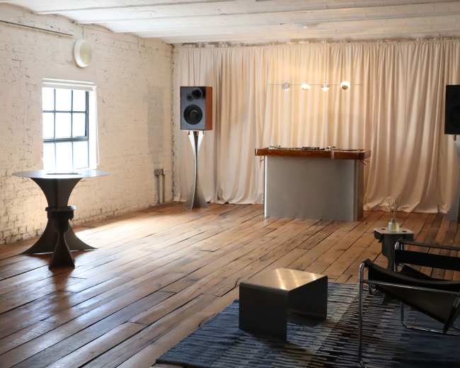 The image shows a minimalist room featuring a wooden floor, a DJ booth with speakers, a small table, and a chair.