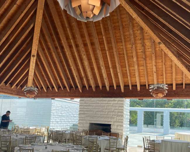 A spacious dining area with round tables set for a meal, featuring wooden beams and modern light fixtures overhead.