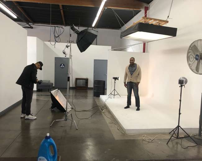 A man is standing on a white backdrop in a photography studio, while another person sets up equipment nearby.