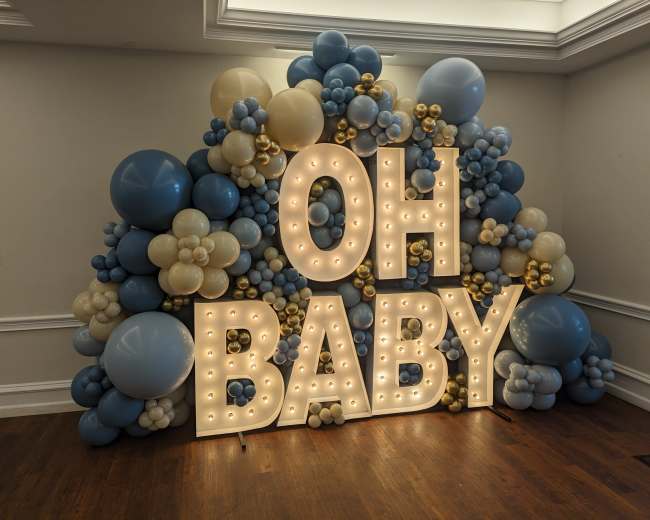 A decorative backdrop made of blue, beige, and gold balloons displays the illuminated phrase "OH BABY."