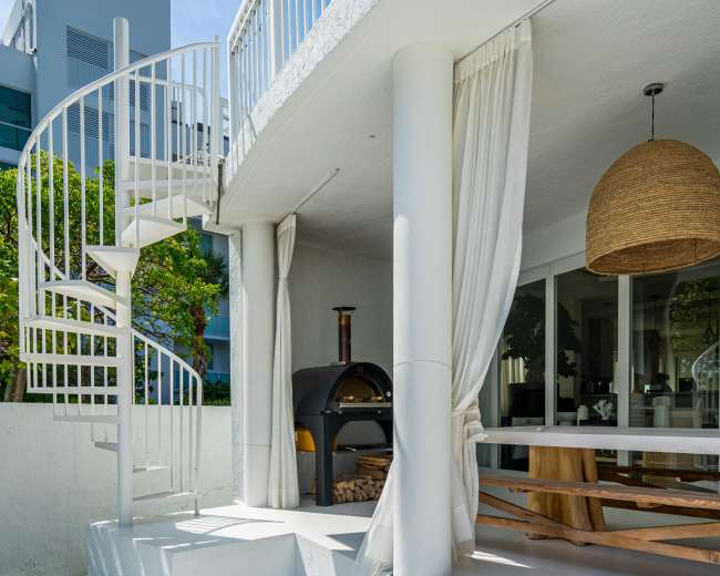 A white spiral staircase leads from a covered outdoor area to a grassy courtyard beside a modern house with an outdoor pizza oven.