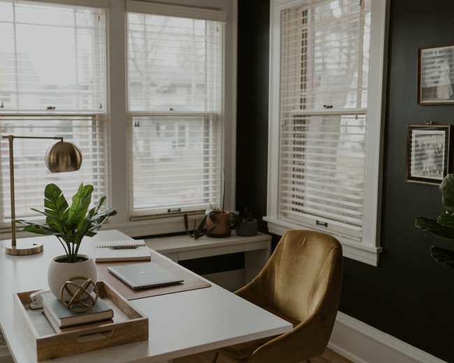A modern office space features a white desk with a laptop, notebook, and decorative items, accompanied by a gold desk lamp and a green plant, set against large windows with blinds.