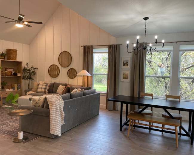 The image shows a cozy living space featuring a gray sofa with decorative pillows, a wooden dining table, a chandelier, and large windows with a view of greenery.