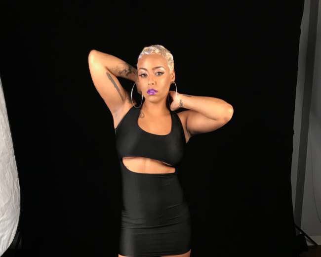 A woman in a black cut-out dress poses with her hands behind her head against a black backdrop.