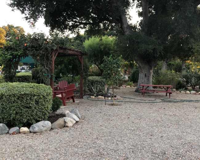 The image shows a landscaped garden with a red wooden arbor, chairs, and a picnic table surrounded by trees and shrubbery.