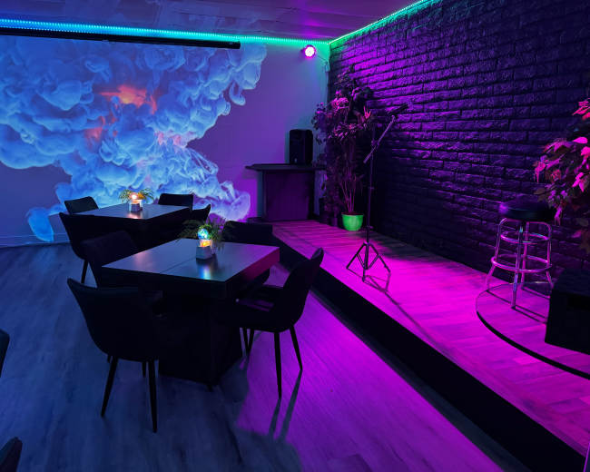 The image shows a modern lounge setting with purple and blue lighting, featuring wooden tables, chairs, and a small stage with a microphone and plants.