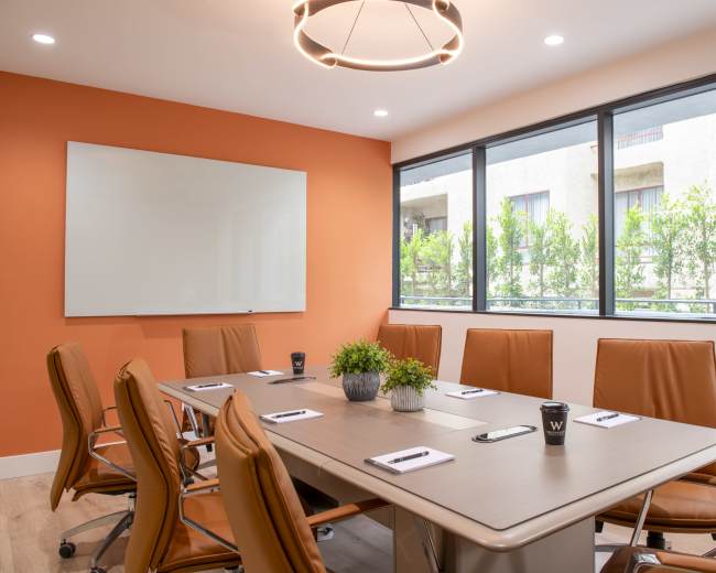 A modern conference room features a large table surrounded by orange leather chairs, with a whiteboard and large windows overlooking greenery.
