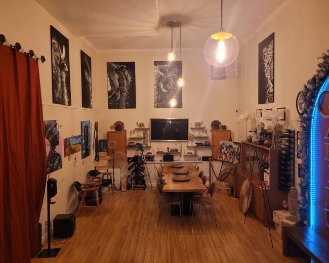 The image shows a spacious art studio featuring wooden furniture, several artworks on the walls, and a central dining table surrounded by chairs.