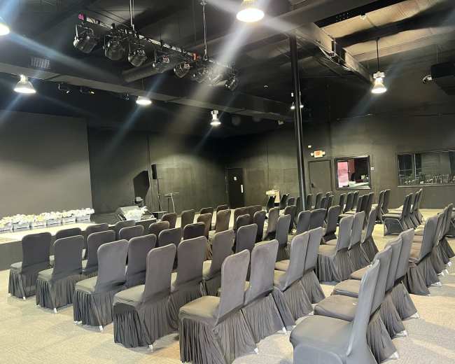 The image shows a spacious indoor venue set up with rows of black upholstered chairs facing a stage area.