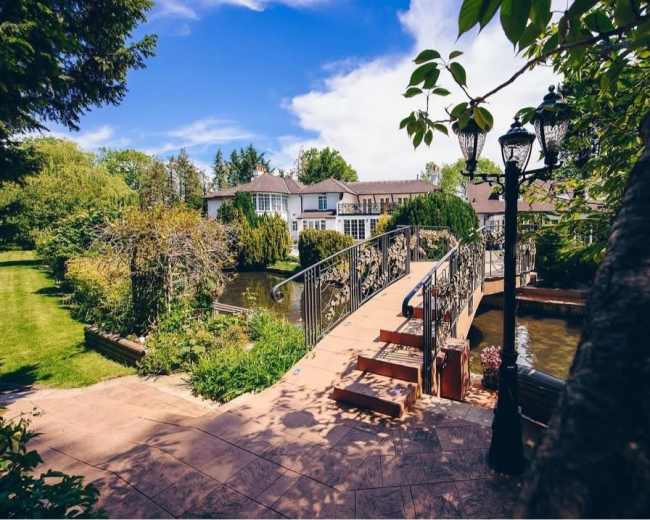 A decorative bridge spans a pond leading to a large house, surrounded by well-manicured gardens and lamps.