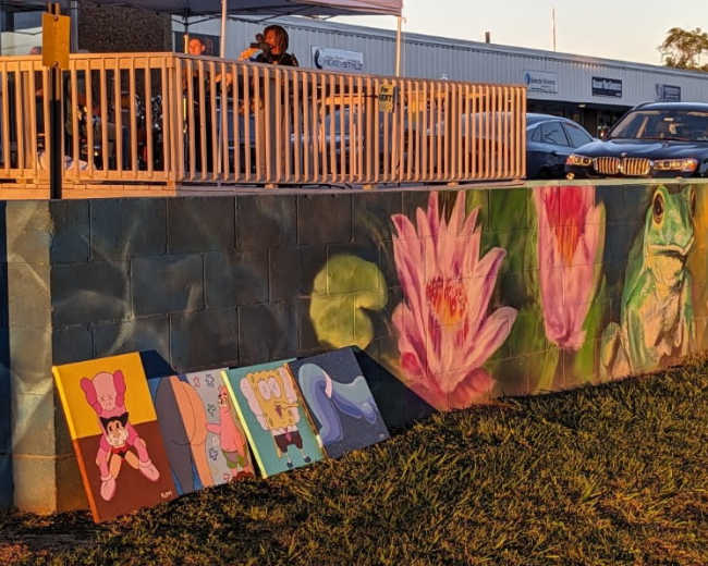 A mural featuring a frog and a water lily on a wall, with several paintings, including a cartoon character, leaning against the wall in front of a deck where people are seated.